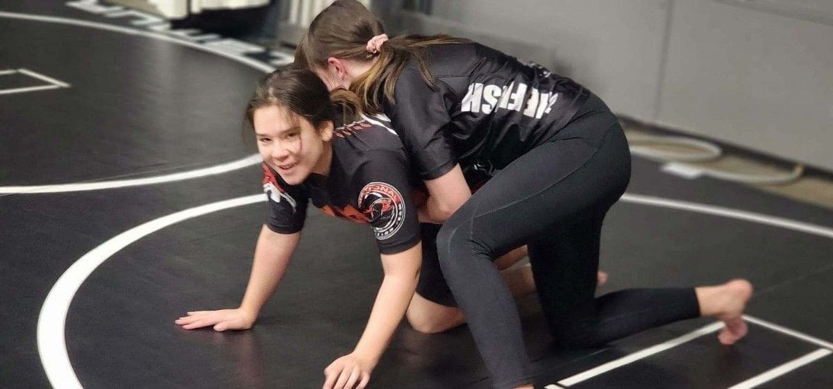 Two girls practicing grappling techniques during brazilian jiu jitsu training on a matted floor.