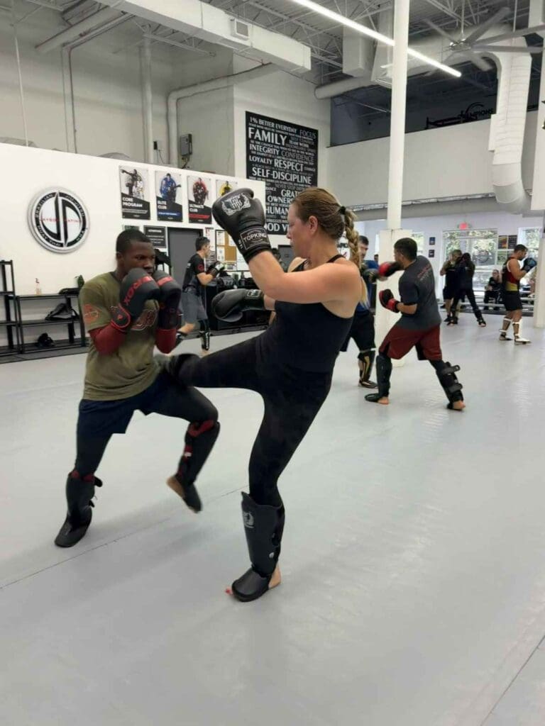 A female kickboxer throwing a push kick toward her sparring partner during an adult training session in a martial arts academy, developing Adults Brazilian Jiu Jitsu position strategy.